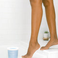 Woman's tanned legs stepping closer to a blue tub of `Sugared + Bronzed` Sugar Paste on top of a tiled floor
