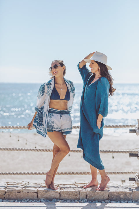 Two women standing on a beach with ocean view, wearing swimsuits and cover-ups.