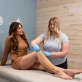 Woman sitting on a bed smiling at the sugarista as she applies and removes sugaring paste from the woman's legs