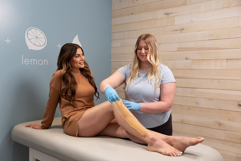 Woman sitting on a bed smiling at the sugarista as she applies and removes sugaring paste from the woman's legs