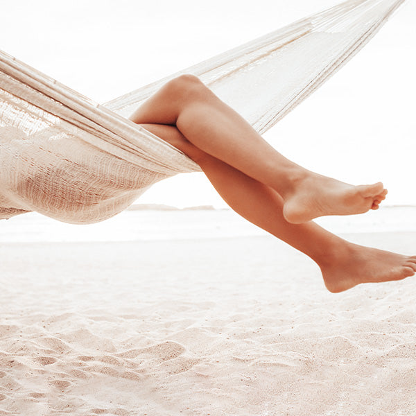 A woman's legs hanging off the edge of a white hammock over white sand