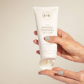 Hand holding a tube of S+B Sunless Tanning Lotion against a beige background