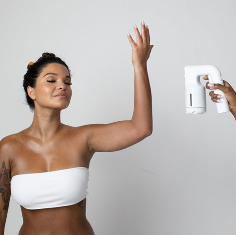 Woman using a white handheld spray tanning device on her arm against a plain background