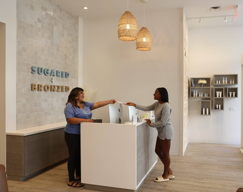 Reception and customer smiling at each other during at transaction at the reception desk in a `Sugared and Bronzed` store lobby.