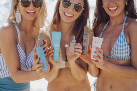 Three women on a beach holding `Sugared + Bronzed` skincare and tanning products.